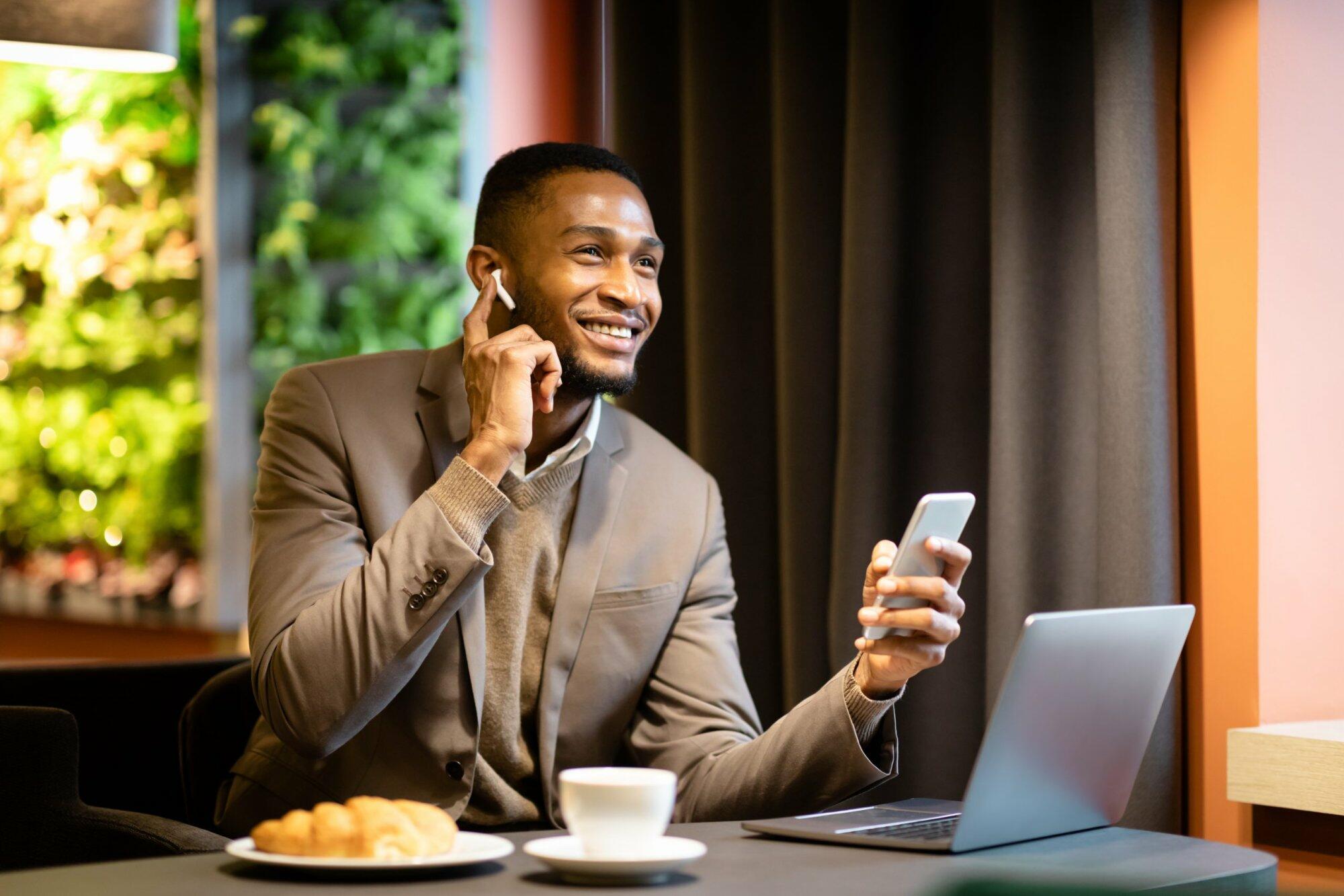 Hands Free Communication. Happy afro businessman speaking on mobile phone in headphones, touching earbud, copyspace