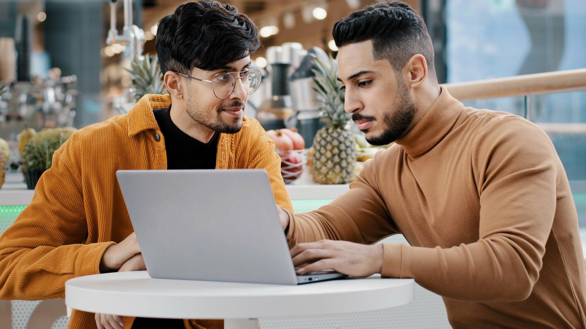 Two arab men sit looking at laptop screen communicate smiles studying using new computer application browsing web pages online dating setting up software young guy explaining to friend helping support. High quality 4k footage